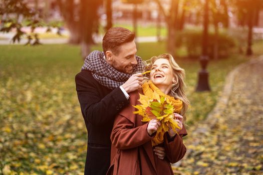 Happy young people in love, man hugging woman from behind stroking her cheek, happy couple walking in a autumn park wearing stylish coats and picking up fallen leaves. Family and people concept,