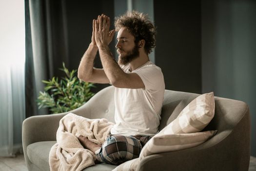 Young Relaxed Man Doing His Daily Yoga on the Sofa in His Apartment. A Man With Praying Hands and Folded Legs Sitting in Lotus Pose. Close-up. High quality photo