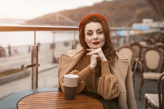 Lovely french young woman sitting at restaurant terrace with coffee mug looking at camera. Portrait of stylish young woman wearing autumn coat and red beret outdoors.