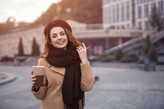 Casual portrait of french woman holding coffee mug outside in autumn beige coat and red beret with urban city background. Tinted photo.