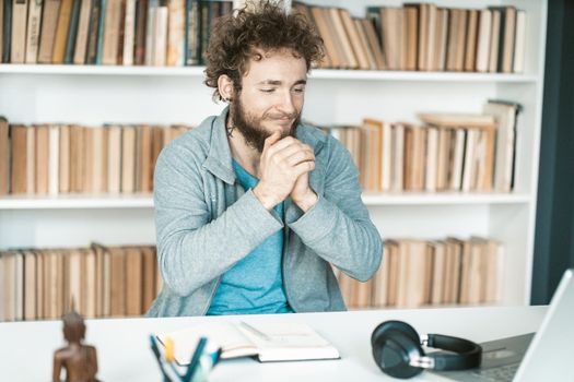 a Resident Gets Nervous Before a Job Interview Online. Excited Man With Folded Hands Sits at a Table in Front of his Laptop. Shelves Full of Books Background. Close-up. High quality photo