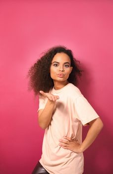 Blowing a kiss african american girl with long curl hair looking at camera wearing peachy t-shirt isolated on dark pink background. Love concept.