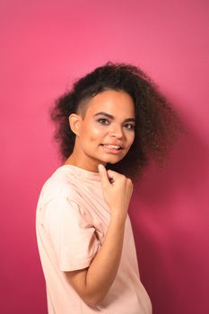 Charming passionate young African American girl with Afro hair looking positively at camera with one arm folded wearing peachy t-shirt isolated on pink background. Beauty concept.