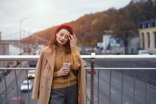 Woman with coffee mug on city street. Charming woman enjoying her cup of coffee standing in a spring urban bridge with traffic behind. French woman in red beret with background of urban city.