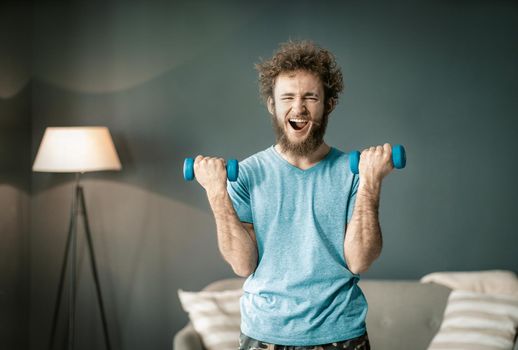 Funny Curly Guy Enjoys Pumping His Biceps by Two Special Dumbbells. Funny Man in blue T-shirt Has Sport At Home. Close -up. High quality photo