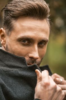 Young man cover his face with a corner of his coat standing on the street in an autumn coat smiling looking at the camera. Handsome young man in dark blue coat and scarf or muffler.