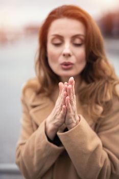 Warming up her hands blowing on the charming young woman standing on the street. Portrait of stylish young woman wearing autumn coat outdoors. Autumn accessories.