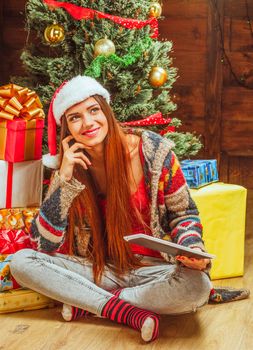 Beautiful Woman in a Winter Knitted Sweater, Hat and Striped Socks Sits on the Floor with Crossed Legs Near a Pile of Gifts and a Christmas Tree. Close-up. High quality photo