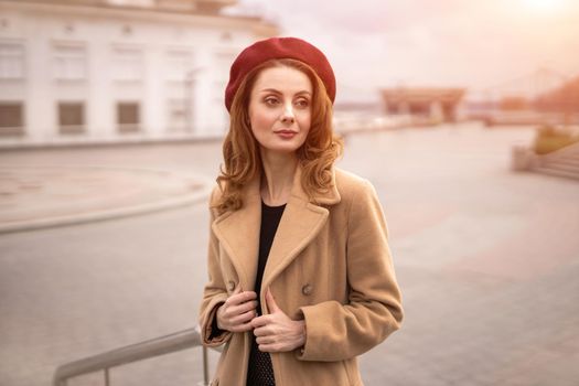 Charming French young woman in an autumn beige coat and red beret standing holding the edges of the coat trying to button it urban city background. Toned photo.
