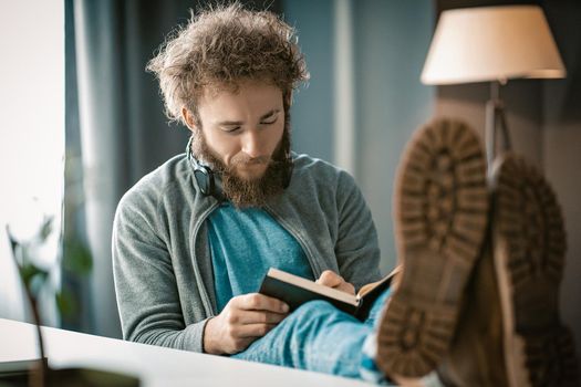 Curly-haired Guy Writes his Thoughts in a Diary. A Man Taking Notes in a Diary. Feet on the Table. Close-up. High quality photo