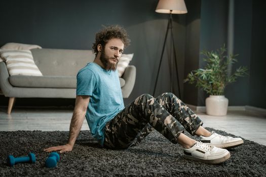 Man in a Blue T-shirt Sits on a Carpet Next to his Blue Dumbbells and Gazes at the Camera. Medium Shot. Living Room Background. High quality photo
