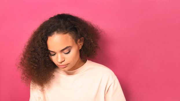 Beautiful but sad African American young girl looking down lowered head wearing peachy t-shirt isolated on pink background. Beauty concept.