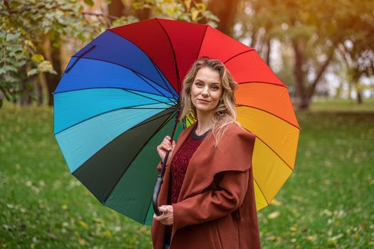 Beautiful young woman standing in the park under a rainbow colored umbrella. A beautiful girl walks through the autumn park in rainy weather.