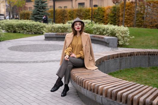 Beautiful young woman in an autumn beige coat and sunglasses sits on a flowerbed bench waiting for her date or girlfriends. Toned photo.