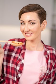 Close up housewife in red plaid shirt and short hair cooking soup on modern kitchen. Smiling woman testing her dish. Chef woman having fun at kitchen.