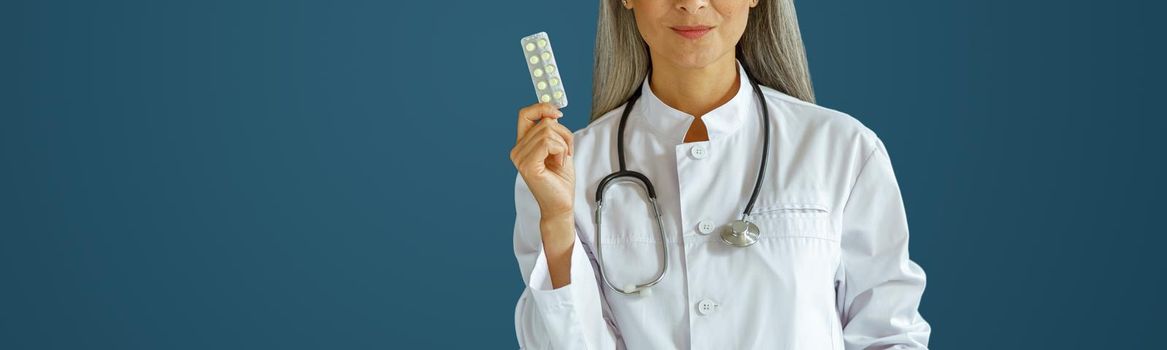 Positive mature woman therapist in white uniform shows pills blister standing on blue background in studio, space for text. Professional medical staff