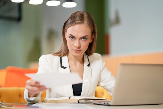 Business Woman in a White Elegant Jacket Sits at her Desk with a Laptop, Gazes to the Camera and Gives Business Papers. Blonde Model Female with Blue Eyes Posing in a Business Jacket in the Office. Burred Background. Close-up. High quality photo