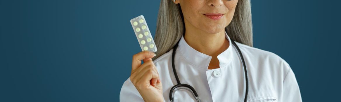 Smiling middle aged female therapist in white uniform holds pills blister standing on blue background in studio. Professional medical staff