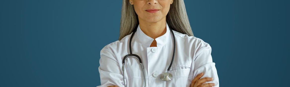 Mature female doctor in white robe with glasses and stethoscope stands on blue background in studio. Professional medical staff