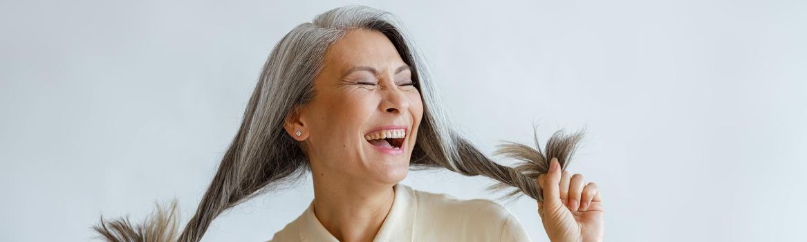 Joyful Asian lady in blouse holds long hoary hair laughing on light grey background in studio. Mature beauty lifestyle