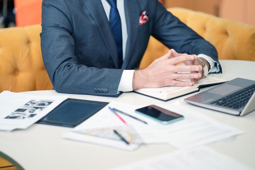 Torso of a Business Man in a Formal Suit, Sitting at his Desk, Hands are on the Diary. On the Table there is a Laptop, Work Papers, Office Supplies, a Smartphone and an Electronic Book. Close-up. High quality photo