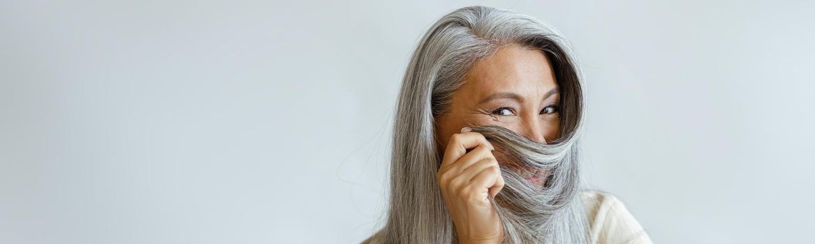 Positive middle aged Asian lady uses hoary hair as niqab sitting on chair on light background in studio, space for text. Mature beauty lifestyle