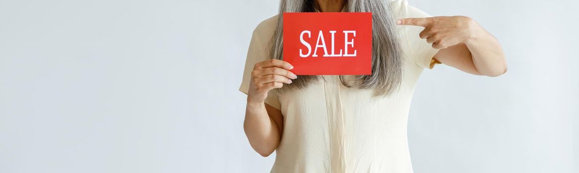 Excited mature Asian woman with long silver hair points onto red Sale sign posing on light background in studio, space for text