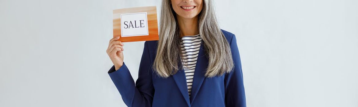 Pretty middle aged Asian lady with long grey hair wearing blue jacket holds card with word Sale posing on light background in studio