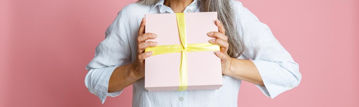 Emotional mature Asian woman with loose grey hair and yellow party hat holds present posing on pink background in studio