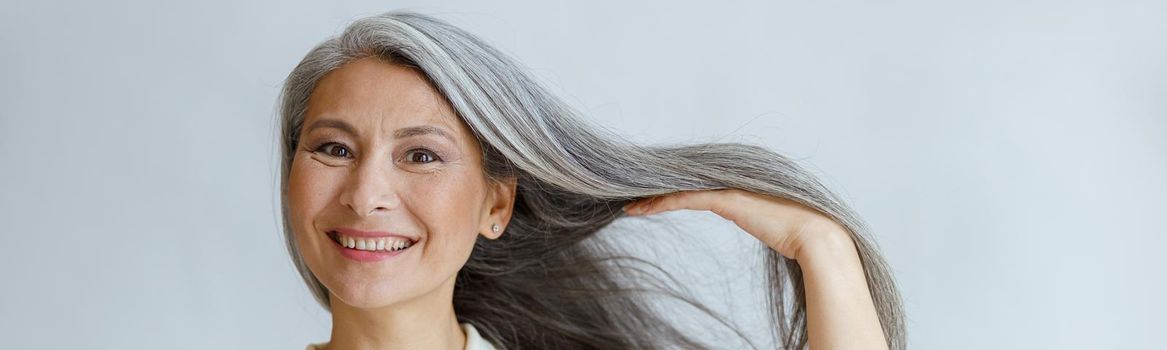 Happy middle aged Asian lady shows natural silver hair posing on light background in studio. Mature beauty lifestyle