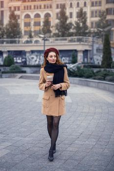 Beautiful french styled young woman with coffee mug on the street female fashion. Portrait of stylish young woman wearing autumn coat and red beret outdoors. Autumn accessories.