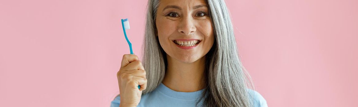 Cheerful middle aged Asian woman with grey hair in blue t-shirt holds toothbrush standing on pink background in studio. Oral cavity hygiene