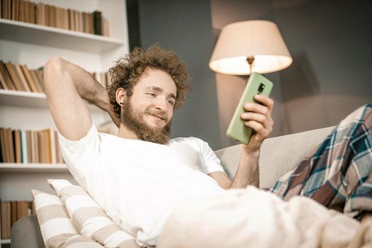 Curly-haired Man Laying on the Couch, Looking at the Phone Screen and Checking Social Media. Romantic Guy Looking for a Girlfriend in Internet. Close-up. High quality photo