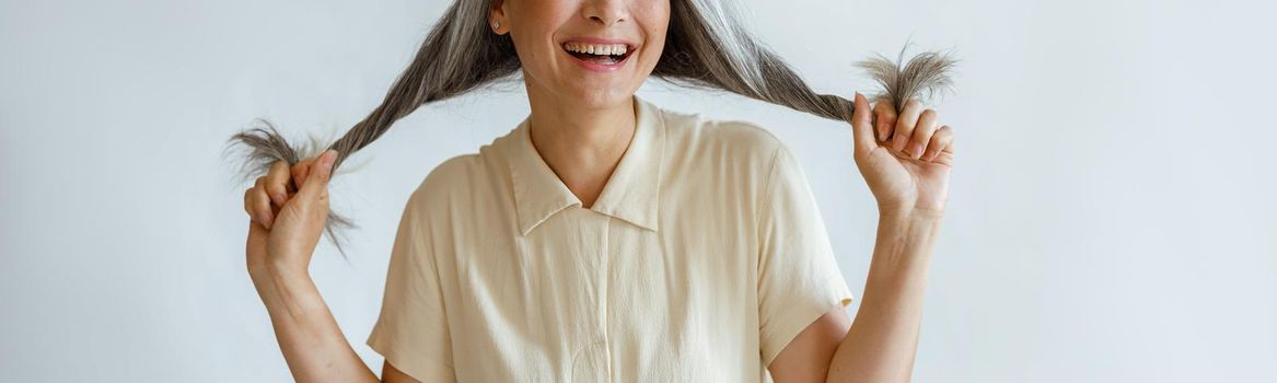 Happy middle aged Asian woman in blouse holds twisted hoary hair smiling on light grey background in studio. Mature beauty lifestyle