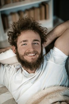 Handsome Guy with Blue Eyes, Curly Hair, a Beard and a Wide Smile Lies Under the Cover in his Bed. Top View. Close-up Portrait. Book Shelves Background. High quality photo