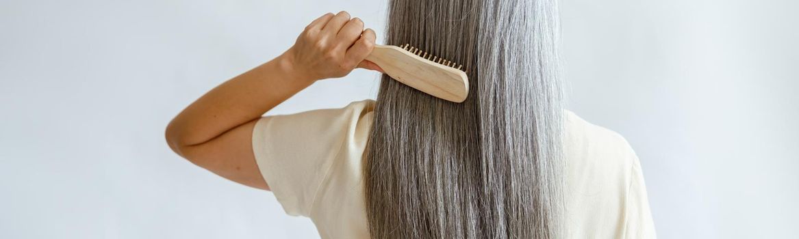 Lady in yellow blouse brushes straight silver hair standing on light grey background in studio backside view. Mature beauty lifestyle