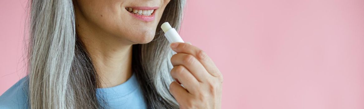 Middle aged grey haired woman in blue t-shirt applies lip balm on pink background in studio closeup, space for text. Mature beauty lifestyle