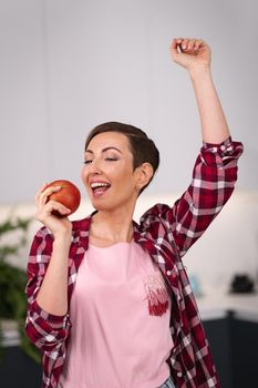 Holding an apple in hand happy woman dancing at kitchen with a sexy eye contact on camera. Charming housewife wearing plaid shirt with a short hair while cooking apple pie standing at the kitchen.