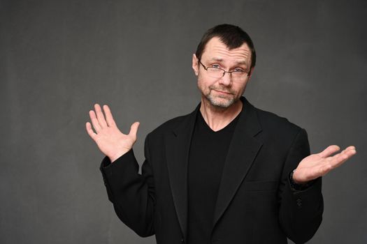 Satisfied man in black jacket with glasses looks at camera with emotion on gray background