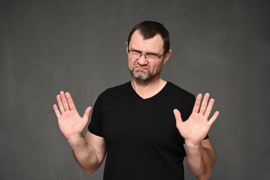 Adult man in a black t-shirt with glasses shows stop with his hands on a gray background