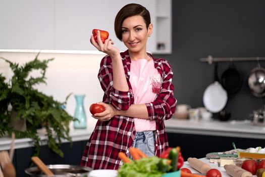 Holding bell pepper and tomato in the hand housewife, wearing plaid shirt with a short hairstyle cooking vegetable ragout at new kitchen in new house. Healthy food at home.
