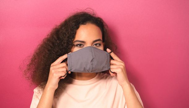 Looking out African American girl covering face with reusable mask in peachy t-shirt, to prevent others from corona COVID-19 and SARS cov 2 infection isolated on dark pink background.