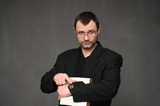 Business man in a black jacket with glasses points to the clock on his hand on a gray background