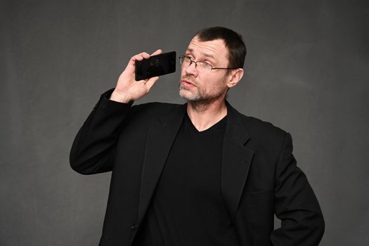 Businessman in a black jacket with glasses talking on the phone with emotions on a gray background