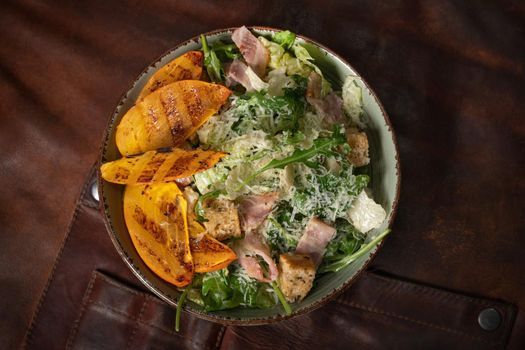 Top view of a bowl of fresh salad with salmon, mozzarella cheese and grilled slices of fruits stands on a table covered with a leather rag. Restaurant concept.