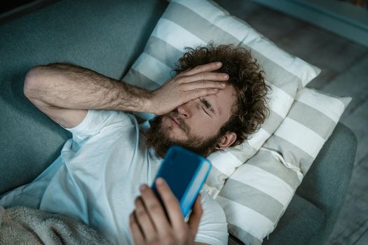 Sleepy Guy Turns Off the Alarm on the Phone. Caucasian Guy Lies on the Bed and Covers Eyes He Has a Smartphone in a Hand. Morning Call, Morning Alarm, Wake up Early. Close-up. Bedroom Background. High quality photo