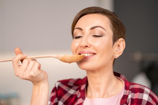 Close-up of a woman trying a cooked dish using a long wooden spoon. Woman with a short hair cooking a dinner for family standing in the modern kitchen of a new house. Healthy food at home.