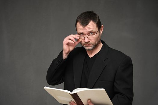 A man in a black jacket with glasses looks at the camera with a folder in his hands on a gray background