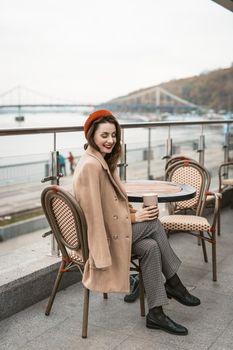 Lovely french young woman sitting at restaurant terrace with coffee mug looking at camera. Portrait of stylish young woman wearing autumn coat and red beret outdoors.