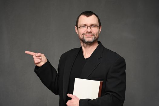 Business man in a black jacket with glasses points to the side with a finger with a smile on a gray background